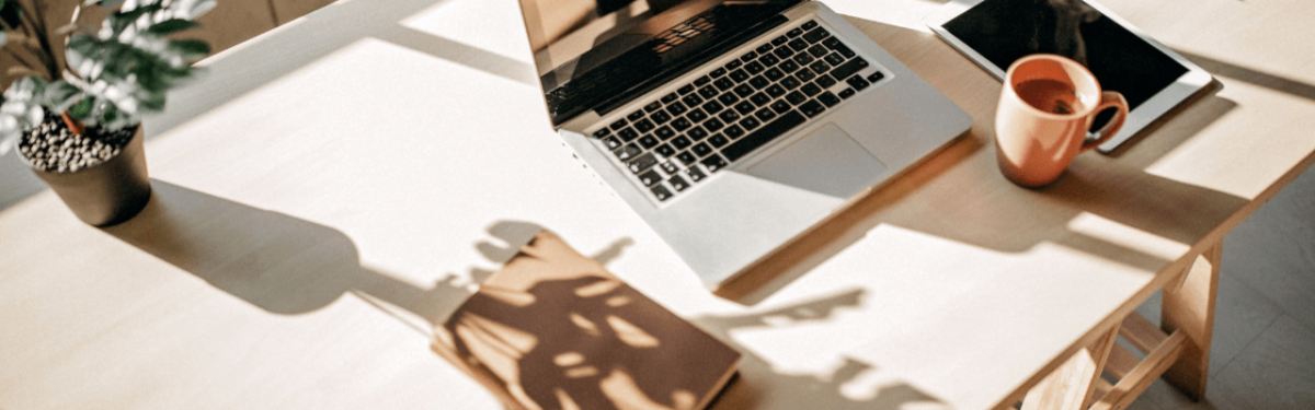 A desk with a laptop, notebook, coffee, tablet and plant on it