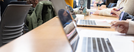 A long desk filled with people taking notes by hand and on laptops